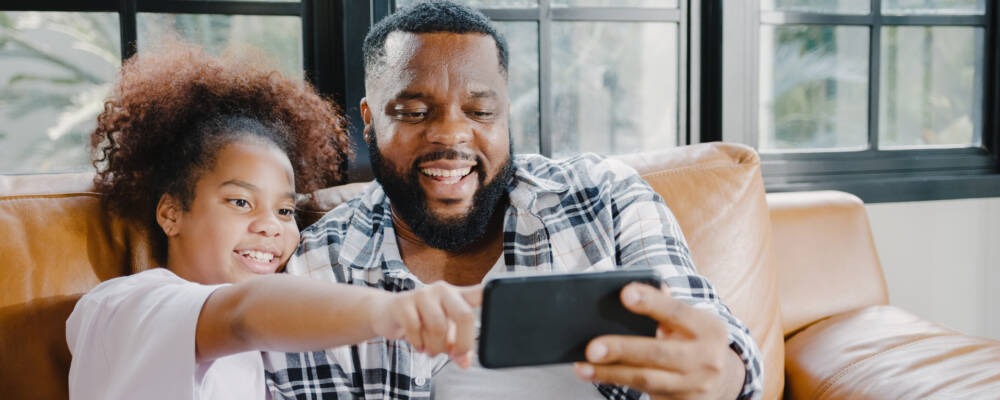 Happy African American family dad and daughter having fun and using mobile phone video call on sofa at house. Self-isolation, stay at home, social distancing, quarantine for coronavirus prevention.